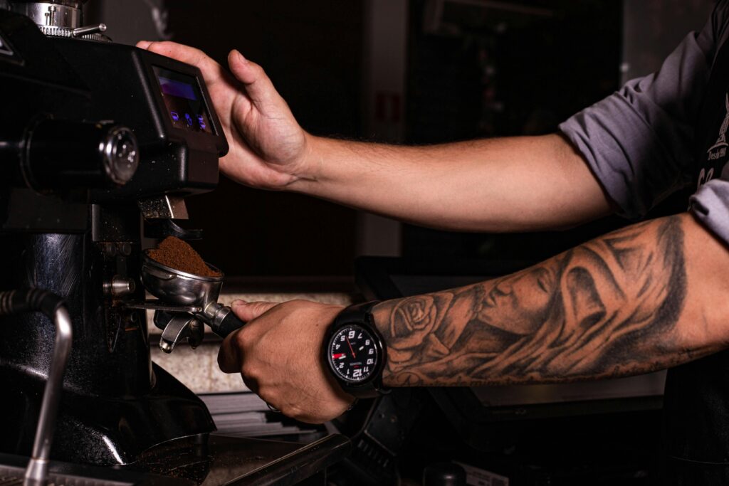 Close-up of a barista's tattooed arm using an espresso machine to grind coffee.